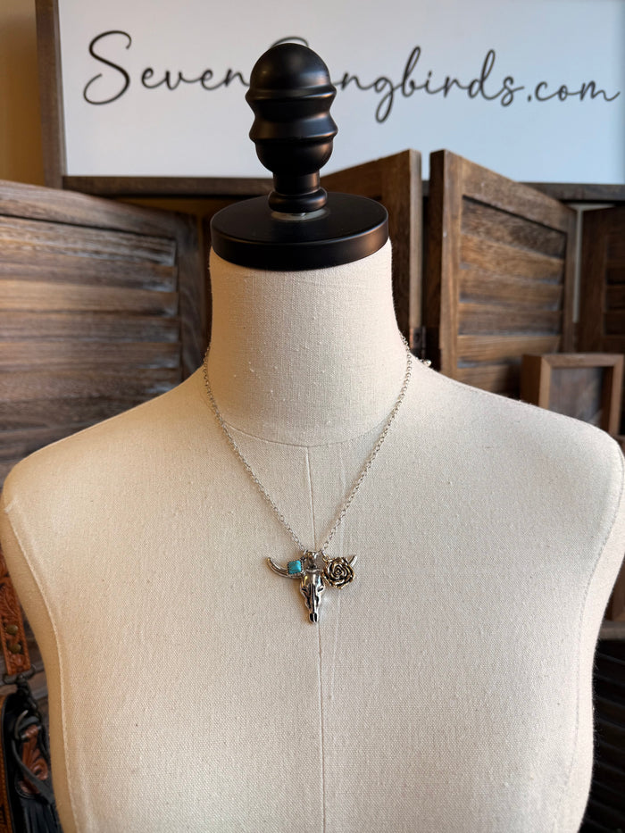 Silver charm necklace with cow skull pendant, blue stone, and rose accent on chain