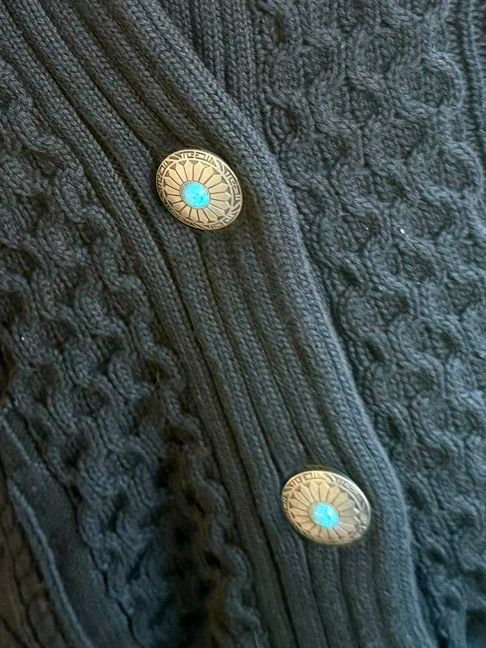Close-up: dark cable-knit fabric with round gold-tone buttons and turquoise centers