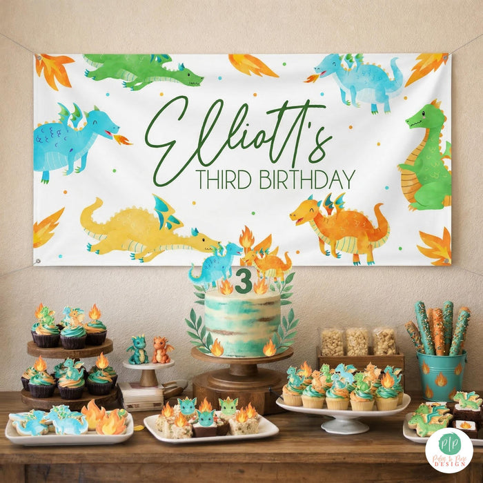 Dragon birthday dessert table, with personalized banner hanging on wall in blue, yellow and green color scheme