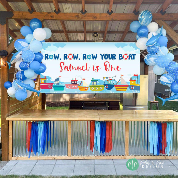 boys first birthday party in a "row your boat" nautical theme with personalized banner displayed in outdoor patio, surrounded by a blue balloon garland, and blue and red streamer decor