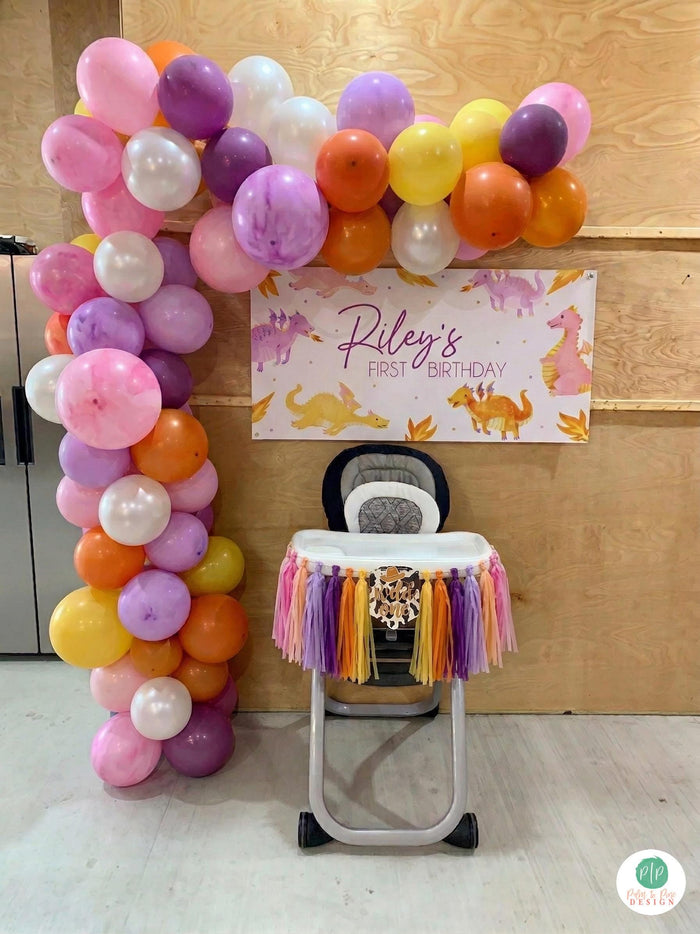dragon first birthday with high chair banner and balloon garland in pink, purple, orange and yellow