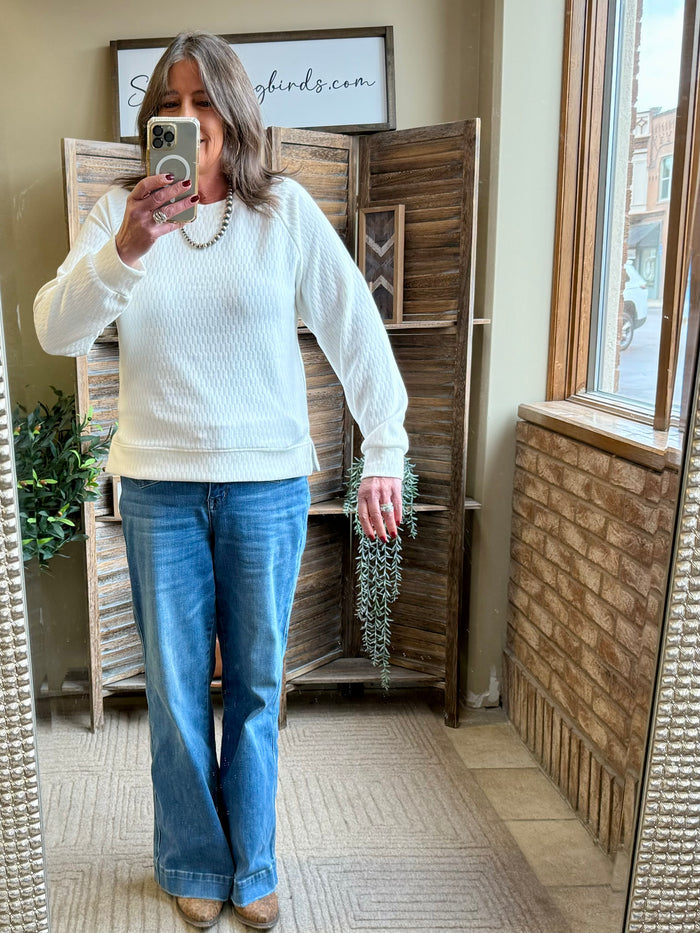 White textured crew neck sweater with long sleeves and ribbed waist, worn with jeans